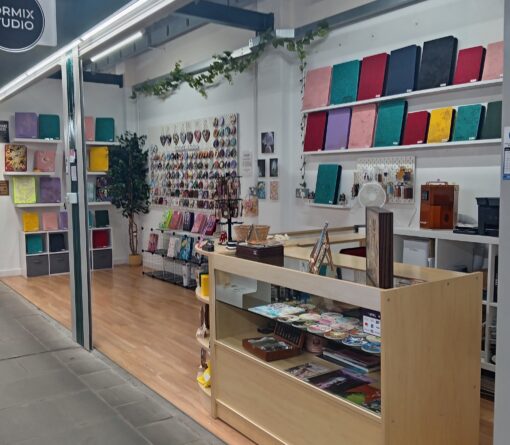 A small art and stationery shop with colourful notebooks on wall shelves, various accessories and items displayed on a central wooden counter, and art prints and keychains arranged neatly on the side wall. The shop has wooden flooring, decorative plants, and bright overhead lighting.