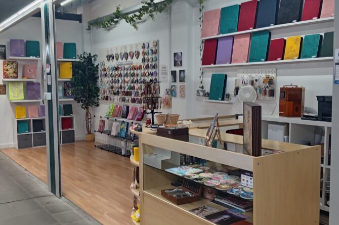 A small art and stationery shop with colourful notebooks on wall shelves, various accessories and items displayed on a central wooden counter, and art prints and keychains arranged neatly on the side wall. The shop has wooden flooring, decorative plants, and bright overhead lighting.